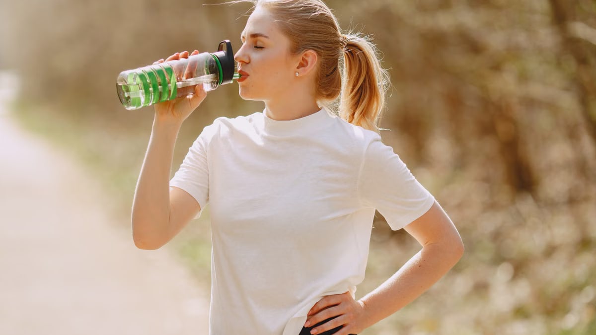 A young woman in a white shirt staying hydrated outdoors with a water bottle in sunlight.